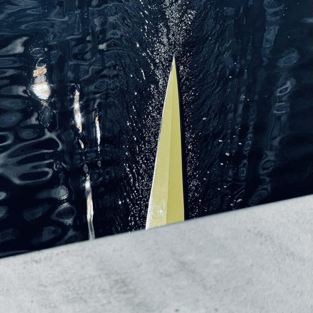 The tip of a rowing boat emerges from beneath a bridge, viewed from above