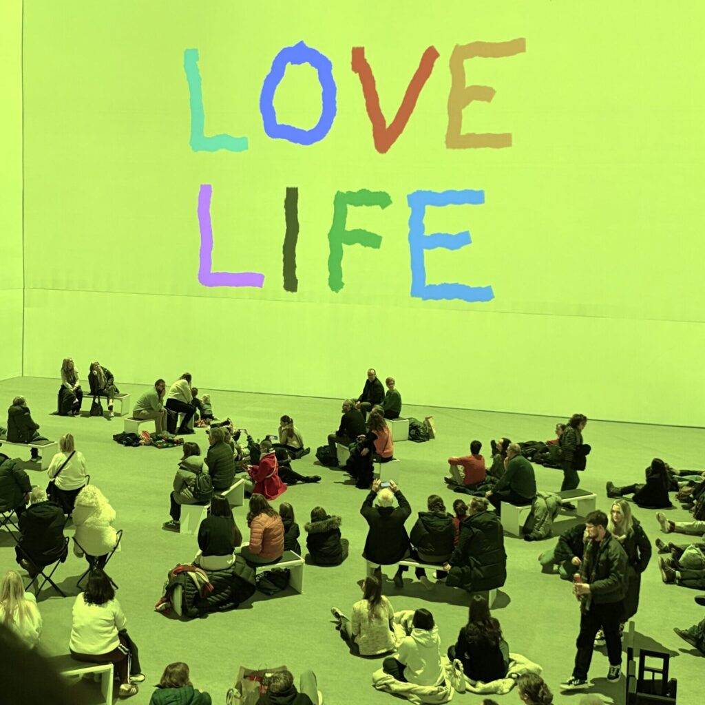 Visitors sit and gather on benches in a large lime-green gallery beneath oversized colorful “LOVE LIFE” lettering. David Hockney Lightroom experience.